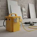 Yellow 110V site transformer standing on a dusty screed floor in a building under construction, with a yellow 110V trailing lead plugged in and running to a corded angle grinder, blockwork wall, plasterboard sheets and a step ladder behind