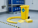 Yellow 120 litre chemical spill kit bin in a factory chemical store, yellow absorbent socks ringing a pale spill on epoxy flooring, blue chemical drums on steel racking and a wall-mounted eyewash station