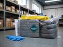 Grey absorbent pads and coiled grey socks from a 120 litre spill kit refill pack wrapped in polythene on a workshop floor, with yellow disposal bags, cable ties, an instruction sheet and blue nitrile gloves