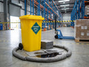 Yellow 120 litre wheeled bin spill kit on a warehouse aisle floor, with grey absorbent socks ringing a dark oil spill and a stack of grey absorbent pads beside it, blue pallet truck and shrink-wrapped pallet behind