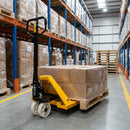 Yellow 2500kg manual hydraulic pallet truck with black tiller handle and white nylon wheels, forks under a shrink-wrapped timber pallet of cartons in a warehouse racking aisle