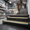 A person walks up a set of black staircase steps with bright yellow grip strips, with two more people behind them in a modern indoor retail or office space with shelves and lighting visible.