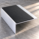A white aluminum L-shaped step with a textured black rubber or foam tread pad, shown on a gray concrete floor.