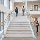 A modern indoor staircase with white steps, glass railings, and light wood handrails, with several people walking up and standing at the top in a bright institutional or office building interior.