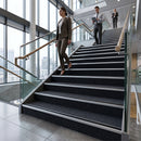 A woman in a brown suit walks down a modern indoor staircase with dark carpeted steps and glass railings, while two men walk further up the stairs in a bright office building.