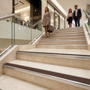 A woman in a beige blazer and black skirt walks down a wide indoor staircase carrying a brown tote bag, with two other people further up the escalator in a modern shopping mall interior.