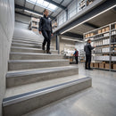 A person walks down a wide set of industrial concrete stairs with integrated metal handrails in a warehouse setting, with shelving and other workers visible in the background.