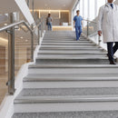 Modern indoor staircase with gray speckled steps and stainless steel handrails, with healthcare workers visible in a bright hospital-like corridor.