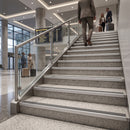 A person in a gray suit walks up a wide indoor staircase with a glass railing in a modern airport or transit terminal, with travelers and luggage visible in the background.