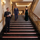 A man in a dark navy uniform stands at the bottom of a grand staircase holding papers, while a man in a blue suit and a woman in a dark green gown walk up the stairs. The staircase has black marble steps with red and white stripes and gold railings.