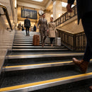 A man in a gray suit and a woman in a beige trench coat walk down a grand hotel-style staircase with black speckled steps, yellow safety stripes, and metal railings, each carrying a suitcase.