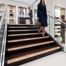 A woman in a navy trench coat walks down a modern staircase in a luxury retail store, holding a brown monogram handbag. The staircase features dark wood steps with brushed silver metal rails and bright orange accent strips.