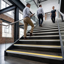 Three people walking up a modern indoor staircase with black steps, silver metal rails, and yellow safety strips. The setting is a bright office or industrial building with brick walls and large windows.