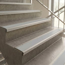 Light gray stair tread strips with integrated metal rails installed on concrete stairs next to a glass railing.