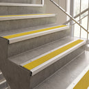 Yellow textured stair tread strips installed on concrete stairs with white metal rails, shown in an indoor stairwell next to a window.