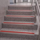 Staircase steps with textured gray concrete-like surfaces and bright red striping between each step, shown indoors next to a metal railing.