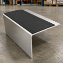 A curved aluminum metal guard rail with a black textured protective mat on top, photographed on a concrete warehouse floor.