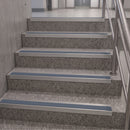 A set of modern stainless steel stair nosing strips installed on gray speckled stone stairs with a metal handrail.