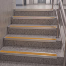 Concrete stairs with textured gray steps and bright yellow tactile striping, bordered by silver metal rails on a staircase landing.
