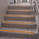 Staircase steps with gray speckled terrazzo-like surfaces, bright orange grip strips, and a stainless steel handrail in an indoor hallway.