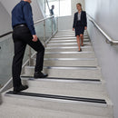 A man and a woman walking up a modern indoor staircase. The stairs have light gray concrete steps with wide silver metal handrails and black grip strips on the steps for safety.