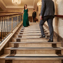 A woman in a floor-length emerald green gown stands on an indoor staircase while a man in a dark gray suit walks up the steps beside her, with a hotel lobby, chandeliers, and marble railings in the background.