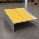 A metal L-bracket shelf riser with two bright yellow textured adhesive pads on the top surface, shown on a concrete warehouse floor.