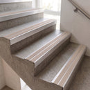 Close-up of an indoor staircase with textured gray concrete steps and beige striping between the steps, alongside a metal handrail against a light wall.