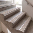 Close-up of an indoor staircase with textured gray stone steps and light beige non-slip rubber tread strips, shown against a white wall with a metal handrail.