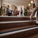 Wooden stair treads with a dark brown wood finish and a silver metal rail, shown in a modern indoor cafe setting with people in the background and a person's legs walking up the stairs in the foreground.