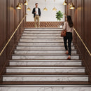 A woman in a beige top and black pants walks up a marble-tiled staircase with dark wood paneling and brass railings, carrying a brown shoulder bag. A man stands at the top of the stairs holding a drink.
