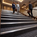 A man in a gray suit and a woman in a blue blazer walk down a grand hotel-style staircase, while a concierge in uniform carries a briefcase up the stairs beside them.