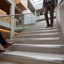 A person in a brown suit walks down wide terrazzo stairs with integrated metal handrails, while another person's legs and a third person are visible in the foreground and background of a modern office lobby.
