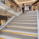 A woman walks up a wide indoor staircase in a modern shopping mall. The steps are light gray speckled stone with bright yellow safety strips, glass railings, and metal handrails.