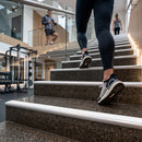 A person in black leggings and gray running shoes ascends an indoor staircase in a modern gym, with another person and exercise equipment visible in the background.