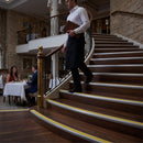 A waiter in a white shirt and dark apron walks down a grand wooden staircase with gold-toned railings in a luxurious restaurant or hotel lobby.