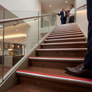 Wood-look stair treads with silver metal rails and red grip strips, shown in a modern indoor staircase setting with people standing at the top and a person's foot in the foreground.