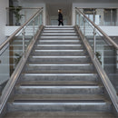 A wide indoor staircase with gray stone steps, silver metal handrails, and glass panels, with a blurred person walking in the distance at the top.