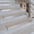 Metal stair nosing strips installed on concrete stairs, featuring a brushed silver metal frame with a beige textured center strip, shown with a stainless steel handrail.