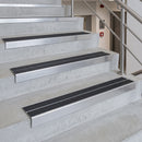 Metal stair nosing strips installed on concrete stairs, featuring a brushed silver aluminum finish with black textured grip strips on the tread surfaces.