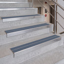 Stainless steel stair tread protectors with dark gray non-slip strips installed on concrete stairs in an indoor stairwell.