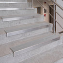 Stainless steel stair tread strips installed on concrete stairs, shown with a metal handrail in an indoor stairwell.