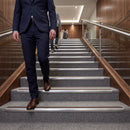 A man in a navy blue suit walks down a modern carpeted staircase with metal handrails, with two other people visible further up the stairs in a corporate hallway.