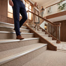 A person in navy trousers and brown shoes walks up a carpeted staircase in a modern indoor setting, with another woman and a man visible further down the stairs in the background.