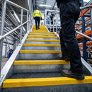 A low-angle view of a person in black work pants and brown work boots climbing a metal staircase with bright yellow safety stripes in a warehouse. Another person in a high-visibility yellow vest is visible further up the stairs.