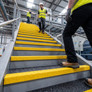 A person in a high-visibility yellow safety vest and black pants walks up a metal staircase with bright yellow handrails inside a large industrial facility. Two other workers are visible further up the stairs.