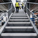 A low-angle view of a person in a high-visibility yellow safety vest and blue hard hat walking up a wide set of industrial metal stairs, with two more workers further ahead in a factory setting.