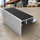 A silver aluminum channel profile with a black textured top surface, shown on a concrete floor in a warehouse setting.