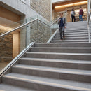 A man walks down a wide indoor staircase with gray stone or tile steps and glass railings. The setting appears to be a modern building lobby or atrium with stone and wood walls, and two other people are visible further up the stairs.