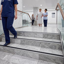 Gray and white marble stair tiles with black metal threshold strips in a modern hospital lobby, with medical staff and a patient visible in the background.